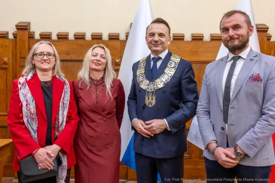 Cztery osoby w formalnych strojach, w tle flagi, uśmiechają się do kamery.