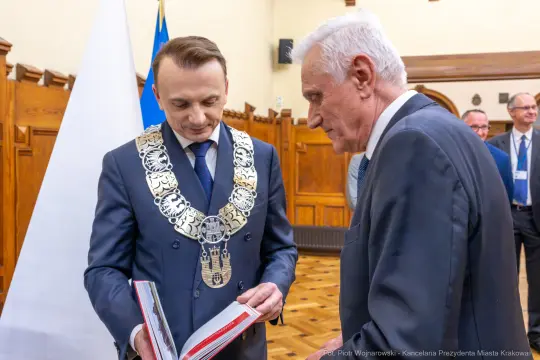 Dwaj mężczyźni w formalnych strojach, jeden z łańcuchem prezydenckim, przeglądają książkę.