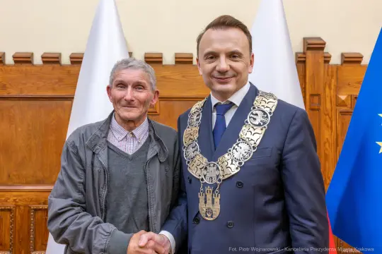 Mężczyźni w garniturach uścisnęli sobie dłonie, w tle flagi i herb.