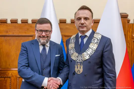 Dwaj mężczyźni w garniturach, jeden z łańcuchem na szyi, uściski dłoni. Flagi w tle.