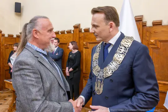 Dwaj mężczyźni w formalnych strojach, uściski dłoni, w tle inni uczestnicy wydarzenia.