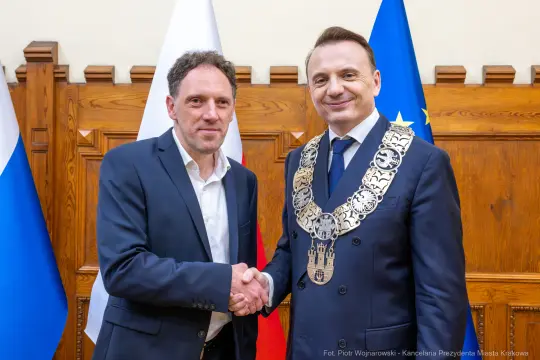 Dwaj mężczyźni uścisnęli sobie dłonie, jeden z nich w łańcuchu prezydenckim. Flagi Polski i Unii Europejskiej