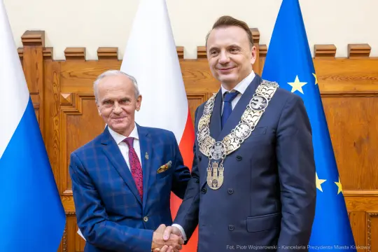 Dwaj mężczyźni w garniturach, jeden z łańcuchem prezydenckim, stoją obok flag Polski i Unii Europejskiej.