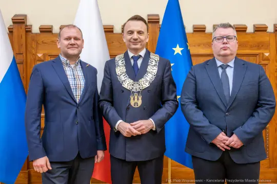 Trzech mężczyzn w garniturach, jeden z łańcuchem prezydenckim, flagi Polski i Unii Europejskiej w tle.