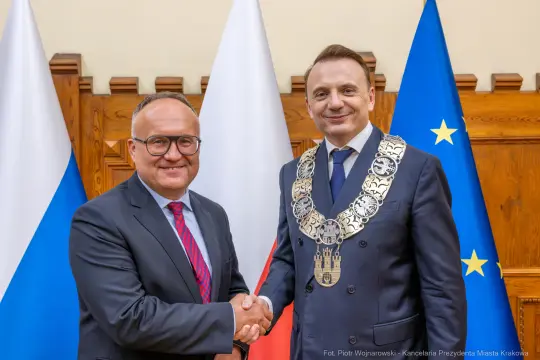 Dwóch mężczyzn w garniturach uścisnęło sobie dłonie, w tle flagi Polski i Unii Europejskiej.