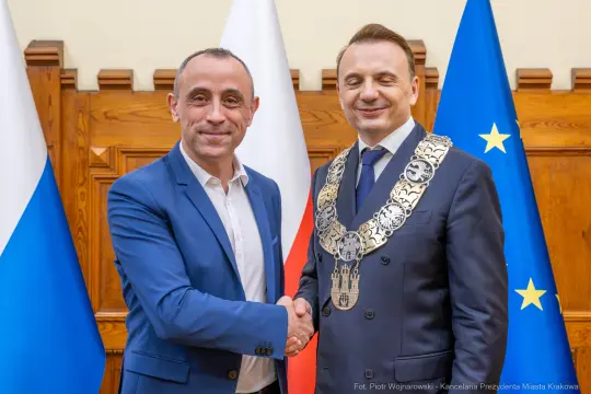 Dwaj mężczyźni w garniturach uścisnęli sobie dłonie, w tle flagi Polski i Unii Europejskiej.