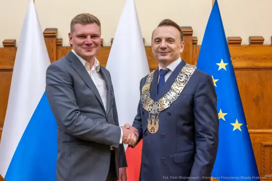 Dwaj mężczyźni w garniturach uścisnęli sobie dłonie, w tle flagi Polski i Unii Europejskiej.