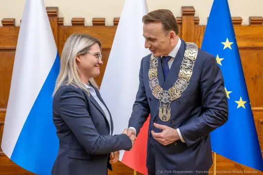 Spotkanie dwóch osób w formalnym uścisku dłoni, w tle flagi Polski i Unii Europejskiej.