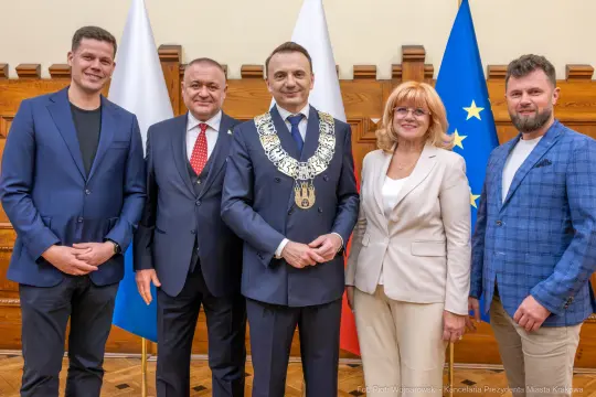 Grupa pięciu osób w formalnych strojach, stojących w sali z flagami.