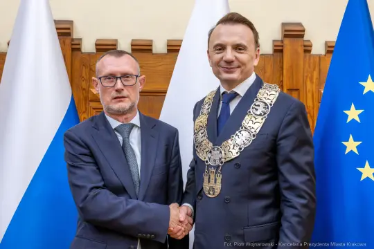 Dwaj mężczyźni w garniturach, jeden z łańcuchem prezydenckim, uściski dłoni, flagi w tle.