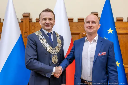 Dwóch mężczyzn w garniturach uścisnęło sobie dłonie, w tle flagi Polski i Unii Europejskiej.