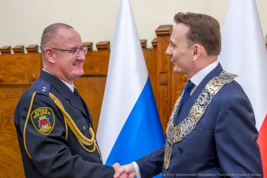 Spotkanie dwóch mężczyzn w mundurze i garniturze, uściski dłoni, flagi w tle.