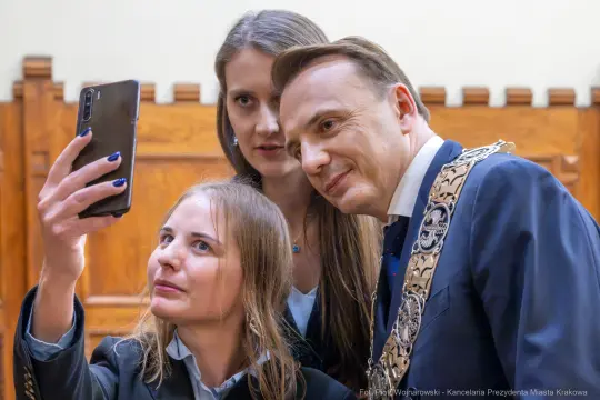 Trzy osoby robią selfie w eleganckich strojach, w tle drewniana ściana.