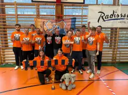 Zespół sportowy w pomarańczowych koszulkach z medalami i pucharem, pozujący na tle flagi.