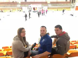 Trzy osoby siedzą na trybunach, w tle odbywają się zawody sportowe.