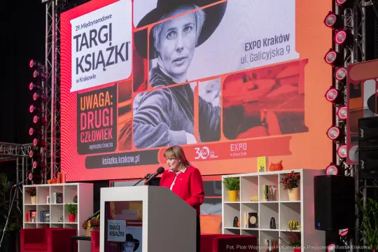 Kobieta przemawia na tle ekranu z informacjami o targach książki w Krakowie.
