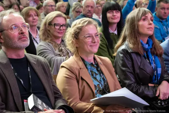 Grupa ludzi na wydarzeniu, niektórzy uśmiechają się, inni słuchają z zainteresowaniem.