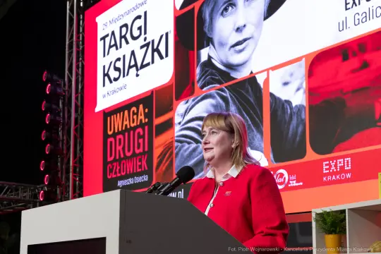 Kobieta w czerwonym ubraniu przemawia na tle plakatu targów książki.
