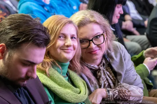 Grupa osób siedzących razem, uśmiechające się kobiety i mężczyzna z telefonem.