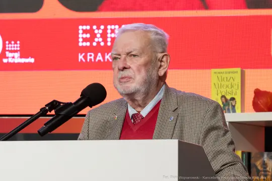 Mężczyzna w garniturze przemawia do mikrofonu na tle kolorowych plansz.
