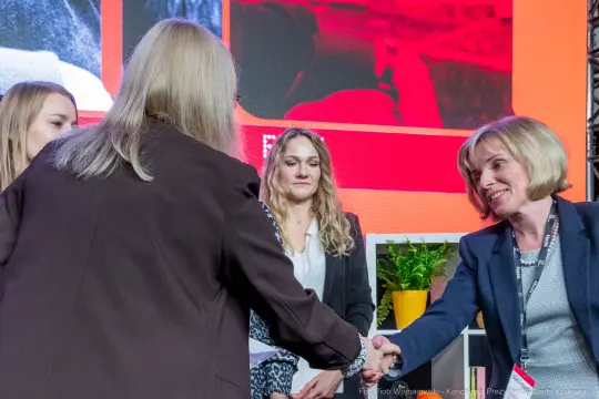 Kobiety na scenie podczas wydarzenia, jedna z nich podaje rękę innej.