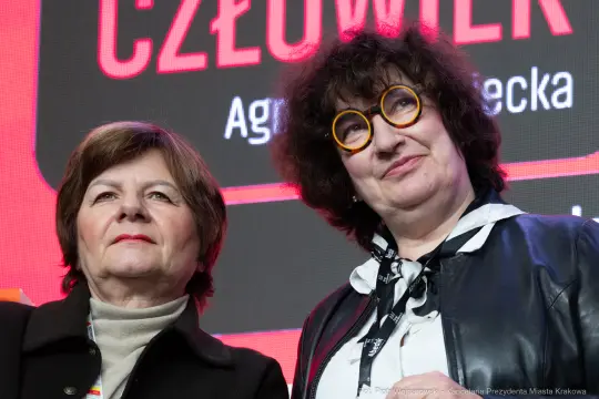 Dwie kobiety z okularami, uśmiechające się na tle kolorowego tła z napisem.