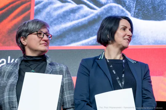 Dwie kobiety na scenie, trzymające kartki, w tle kolorowe tło.