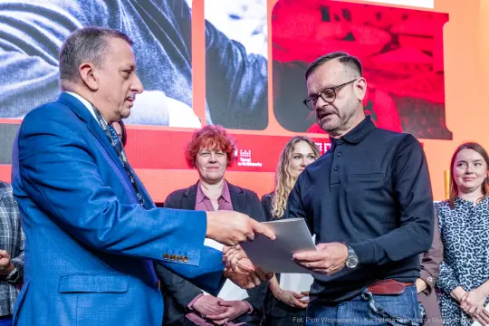 Wręczenie nagrody przez mężczyznę w garniturze drugiemu mężczyźnie w okularach. W tle widoczni inni uczestnicy.