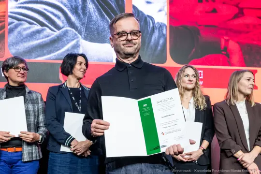 Mężczyzna trzymający dyplom nagrody, w tle grupa kobiet.