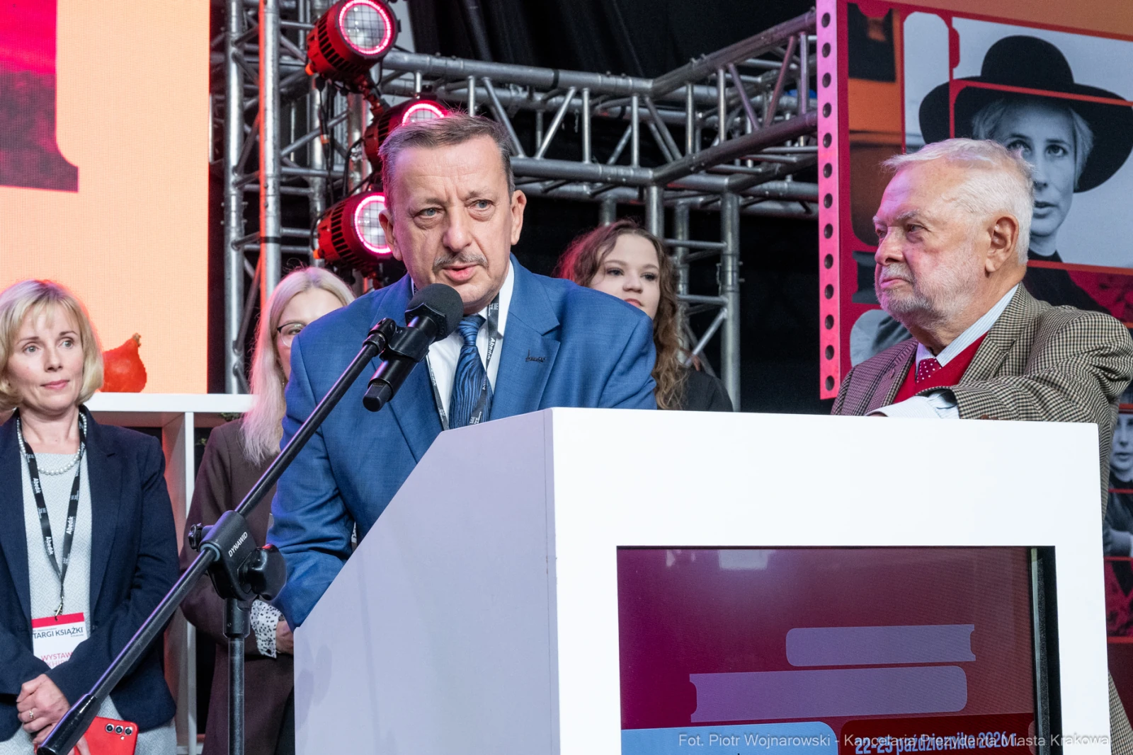 Mężczyzna w garniturze przemawia do mikrofonu, obok stoi starszy mężczyzna. W tle widoczne osoby i dekoracje.  Autor: Piotr Wojnarowski