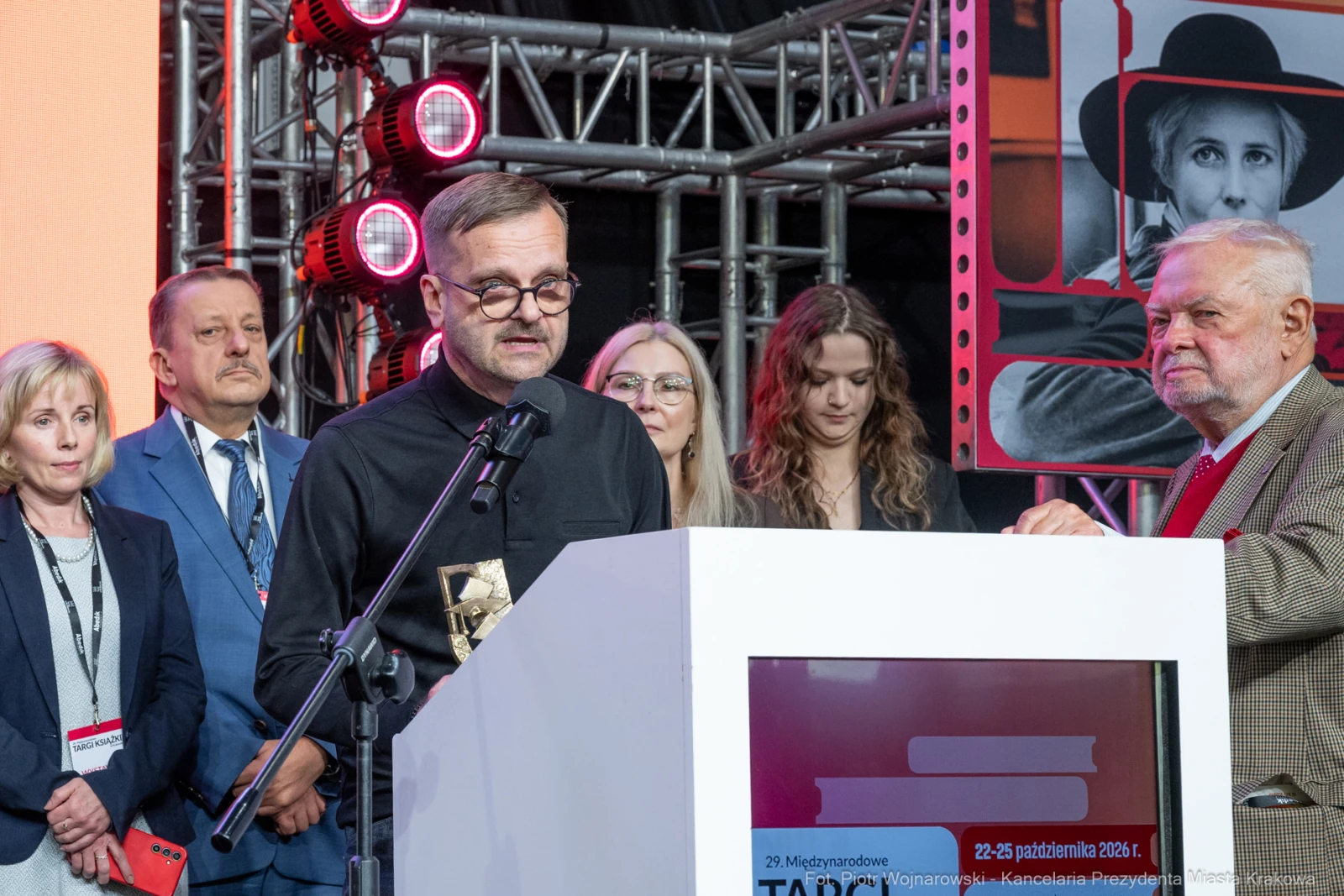 Mężczyzna przemawia na scenie, obok niego stoją inne osoby, w tle widoczne elementy dekoracyjne.  Autor: Piotr Wojnarowski