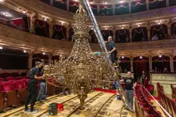 Pracownicy konserwują żyrandol w teatrze, używając drabiny i narzędzi.