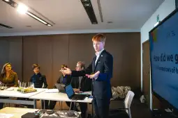 Mężczyzna w garniturze prezentuje przed grupą ludzi w sali konferencyjnej.