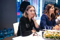 Kobieta w czarnym żakiecie siedzi przy stole, obok innych osób, na stole jedzenie i napoje.