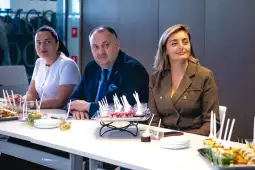 Trzy osoby siedzą przy stole z jedzeniem, skupione na rozmowie.