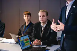Dwóch mężczyzn siedzących przy stole, jeden z laptopem, drugi słuchający prezentacji.
