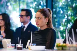 Kobieta w czarnej bluzce siedzi przy stole, skupiona na wydarzeniu, w tle inni uczestnicy.