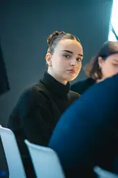 Młoda kobieta w czarnym swetrze, siedząca na tle ciemnej ściany.