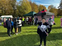 Uczestnicy wydarzenia sportowego w parku, stoiska z informacjami i osoba z numerem startowym.