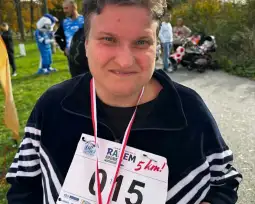 Kobieta z medalem na szyi, uśmiechająca się po biegu na 5 km. W tle widać innych uczestników i dzieci w wózkach