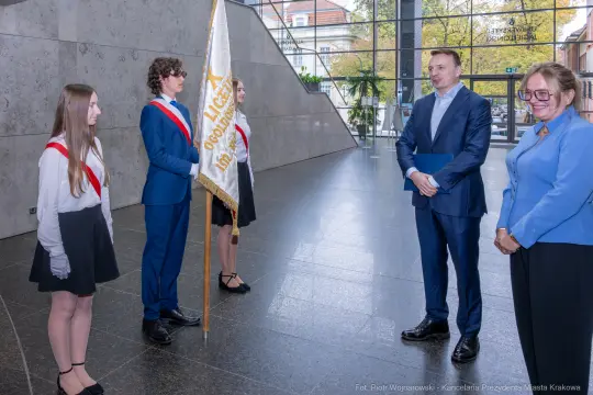 Ceremonia wręczenia flagi w nowoczesnym wnętrzu budynku, uczestnicy w strojach galowych.