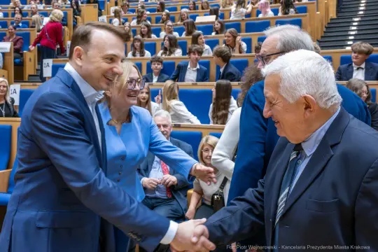 Spotkanie osób w sali, uściski dłoni i uśmiechy. W tle młodzież siedząca na widowni.