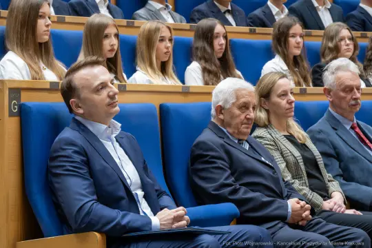 Grupa ludzi siedzących na sali, wśród nich mężczyźni w garniturach i kobiety w białych bluzkach.
