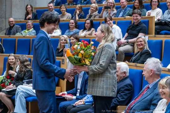 Mężczyzna wręcza kobietę bukiet kwiatów w trakcie uroczystości. W tle zgromadzeni goście.