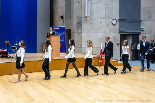 Uczniowie w strojach galowych idą w kierunku podium podczas uroczystości szkolnej.