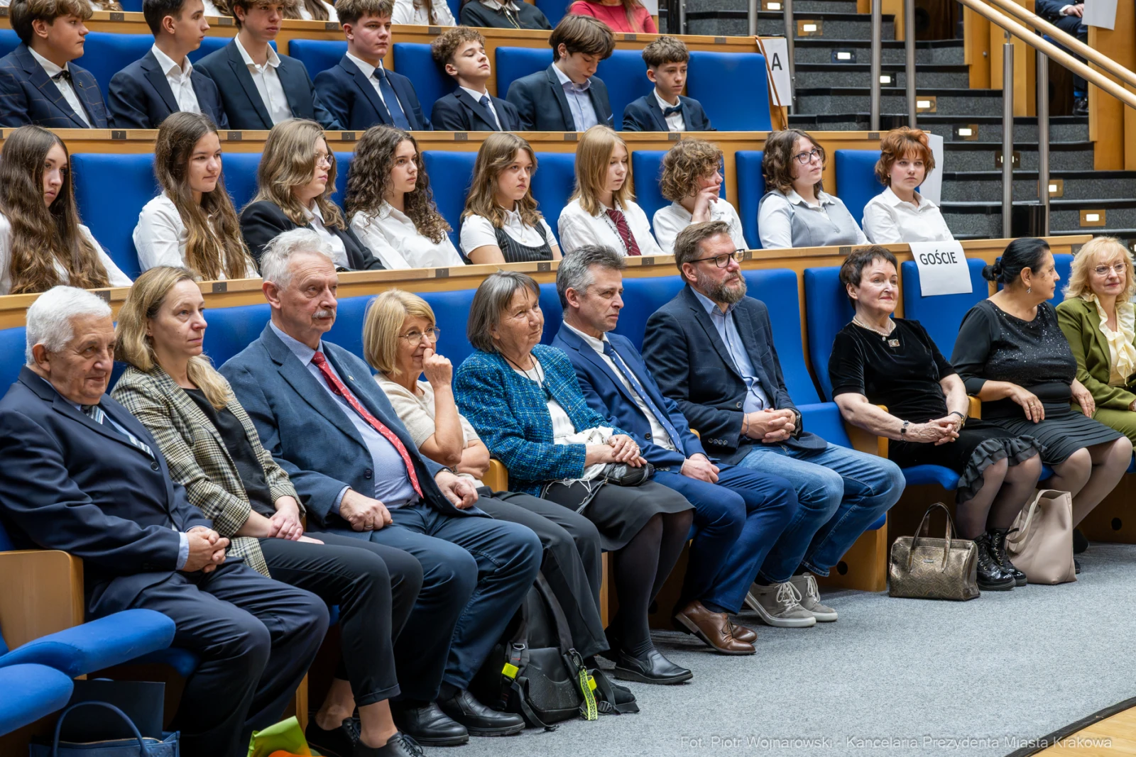 Grupa osób siedzących na widowni podczas wydarzenia, w tle młodzież w strojach formalnych.  Autor: Piotr Wojnarowski