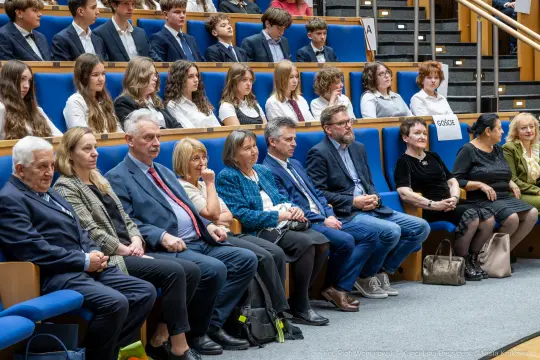 Grupa osób siedzących na widowni podczas wydarzenia, w tle młodzież w strojach formalnych.