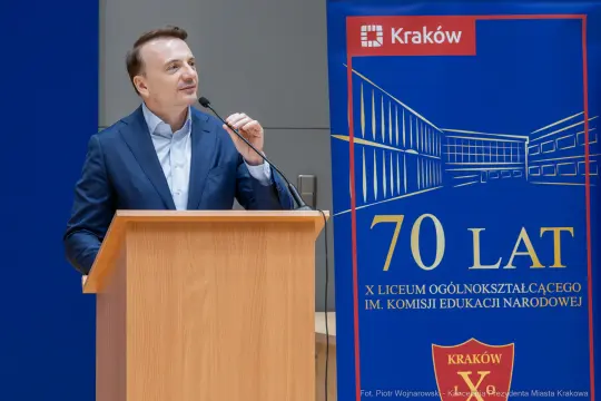 Mężczyzna przemawia przy mównicy obok banneru z okazji 70-lecia liceum w Krakowie.