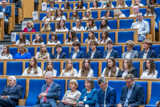 Grupa uczniów i dorosłych siedzących na widowni w sali.