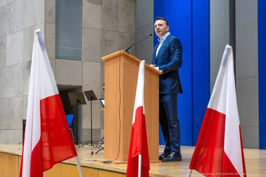 Mężczyzna przemawia z mównicy, obok flagi Polski.